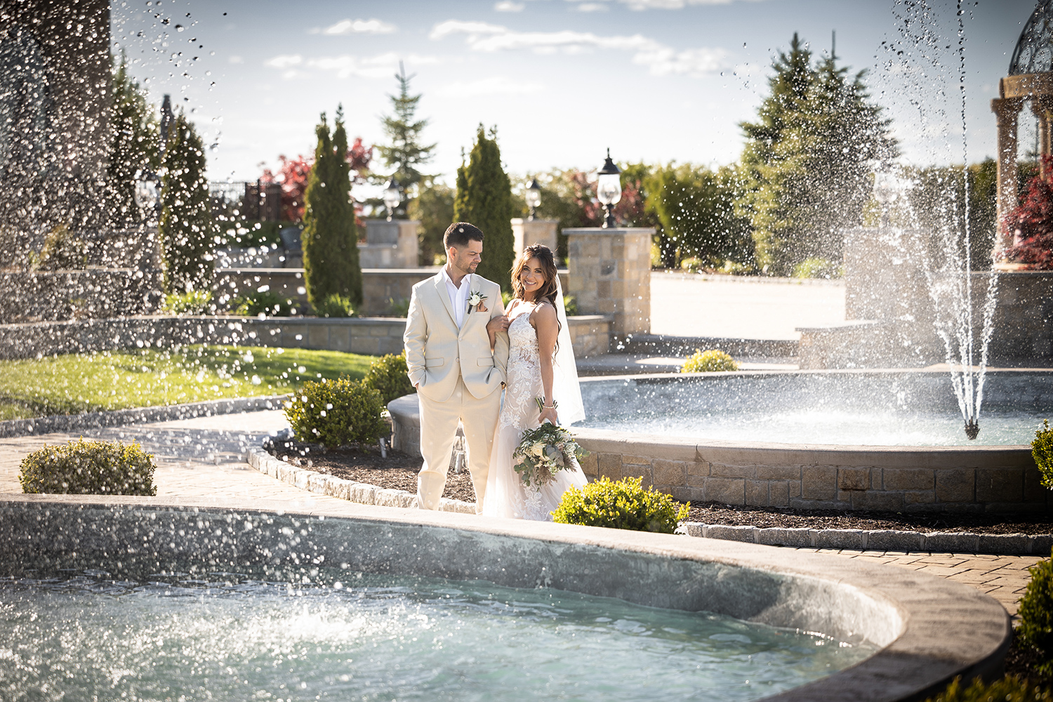 a bride and groom pose behind a fountain outside while the groom looks at the bride