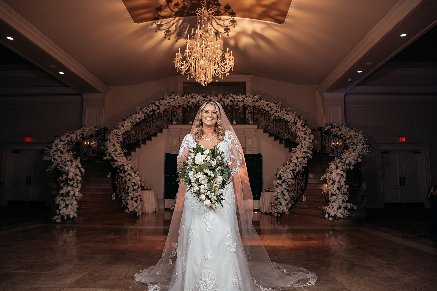 a bride standing in the aria foyer holding a bouquet of white flowers. the railings of the stairs are fully decorated in white flowers
