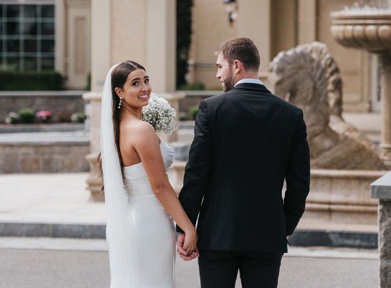 Bride and groom holding hands outside while the bride looks back at the camera and smiles